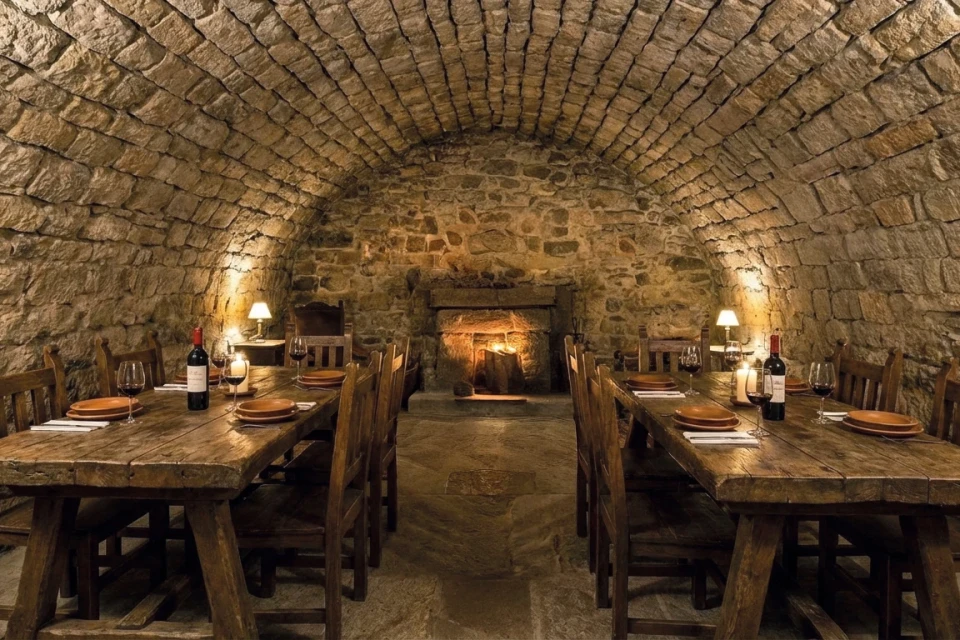 Rustikaler Gew&ouml;lbekeller aus grobem Naturstein mit zwei langen Holztischen und je sechs Holzst&uuml;hlen pro Seite. Auf den Tischen braune Teller, wei&szlig;e Stoffservietten, Weingl&auml;ser mit rotem Wein, zwei Weinflaschen und Kerzen. Seitlich zwei kleine Tischlampen mit warmem Licht. Am Ende des Raums steinerner Kamin mit glimmendem Feuer, Boden aus Steinplatten, Perspektive zentral auf den Kamin ausgerichtet.