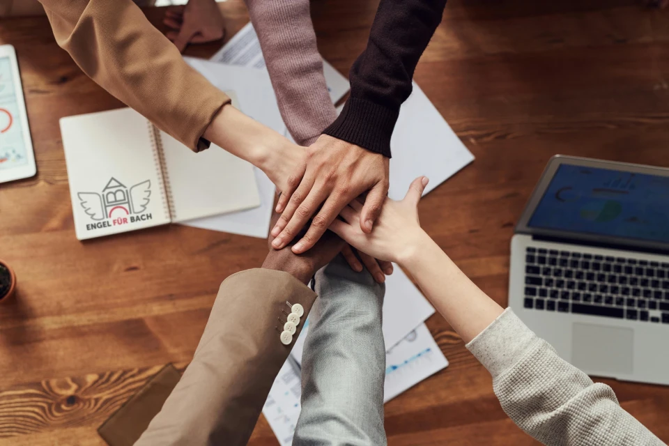 Fünf Menschen strecken ineinander die Hand aus. Sie sind bereit mit der Arbeit zu beginnen.